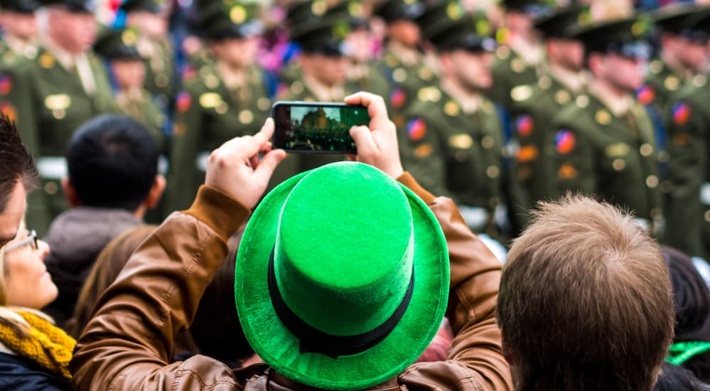 A man in a St. Patrick's Day hat is photographing a parade on his phone near Dayton, OH.